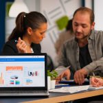 Business woman talking about financial project, taking notes, discussing start up ideas using laptop. Diverse employees gathered in co-working, working process in busy company, teamwork help concept